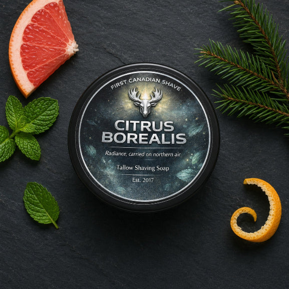 Overhead shot of a jar of Citrus Borealis Tallow Shaving Soap by First Canadian Shave, on a dark stone surface, styled with a sliced grapefruit, mint leaves, pine branches, and orange peel slices.
