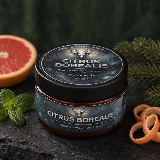A jar of First Canadian Shave, Citrus Borealis Tallow Shaving Soap on a dark stone surface, styled with a sliced grapefruit, mint leaves, pine branches, and orange peel slices.