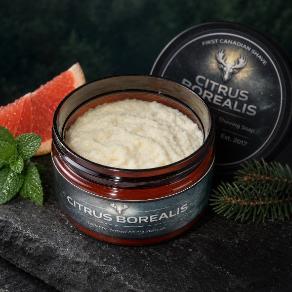 An open jar of Citrus Borealis Tallow Shaving Soap by First Canadian Shave on a dark stone surface, styled with a sliced grapefruit, mint leaves, pine branches, and orange peel slices.